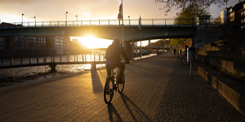 Man riding a bike at the Weser promenade in Bremen, Germany at sunset with long shadows and the sun beaming below a bridge - obrazy, fototapety, plakaty