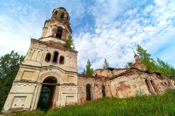 Fototapeta na wymiar View of the front side of an old, destroyed and abandoned church in Russia. Walls with peeling paint and old red bricks. Trees on the roofs. Summer. Daylight. Sky with clouds.
