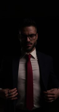 dramatic bearded businessman with glasses fixing and closing suit, holding hands and fading away in the dark