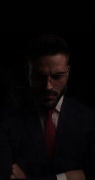 elegant bearded businessman in suit with metal frame glasses crossing arms, smoking and looking to side in the dark