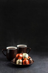 Chocolate candies wrapped in multicolored foil and two cups of coffee on black background