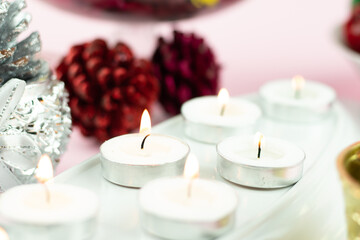 Obraz na płótnie Canvas White Tealight Candles Illuminated In Row With Maroon Pine Cone Glitter Decorations On Light Pink Background. Theme For Merry Christmas And Happy New Year