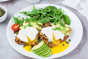 Healthy breakfast. Poached eggs on toast with avocado pieces, arugula, mizuna and chard leaves and cherry tomatoes on a plate on a served table. Flexetarian diet. Close-up