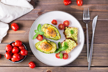 Ready-to-eat appetizer baked avocado with egg, toast and cherry tomatoes on a plate on a wooden table. Healthy eating. Flexitarian diet. Top view