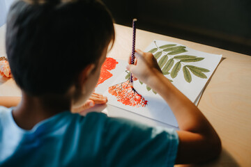 Cute child siting at desk and drawing rowanberries on album sheet with dry rowan leaves