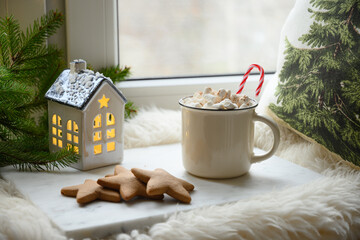 Winter coffee with marshmallow on windowsill with pillow and fur skin decorated fir branches and little toy house. Xmas. Christmas mood.