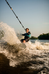 Athletic guy holds rope and speedly riding wakesurf board on splashing wave.