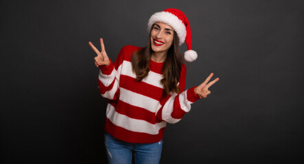 Excited beautiful smiling happy young woman in Christmas Santa hat shows the Victory sign and looks on camera