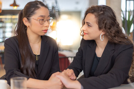 Dos mujeres de negocios, asi&aacute;tica y cauc&aacute;sica, se miran y se cogen las manos. Cauc&aacute;sica sonriente y asi&aacute;tica seria