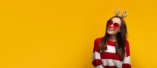 Banner wide photo of an attractive excited smiling young girl in Christmas reindeer antlers and sweater while she posing isolated on yellow background in studio