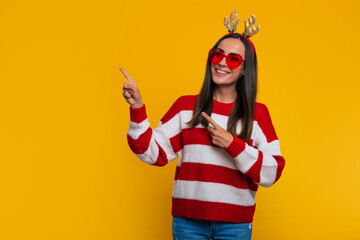 Close up portrait of an attractive excited smiling young girl in Christmas reindeer antlers and sweater while she pointing away on yellow background in studio