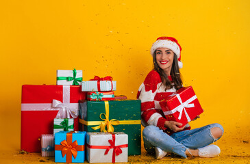Full length photo of excited young smiling beautiful and charming woman in Santa hat and with Christmas mood while she sitting with many colorful gift boxes and having fun with confetti