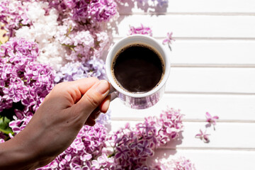Obraz na płótnie Canvas coffee mug in hand.Hands of young woman drinking coffee on white wooden table with flowers, gift. Woman hand hold cup of black coffee, spring flowers, lilac bush. Spring mood. Beautiful breakfast