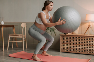 Pregnant woman exercising with fit ball at home, making squats with fitball.