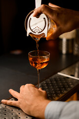 hand of bartender carefully holds strainer on mixing cup and pours liquid into wine glass