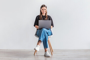 Happy young woman working online, sitting on chair and using laptop against white studio wall, full...