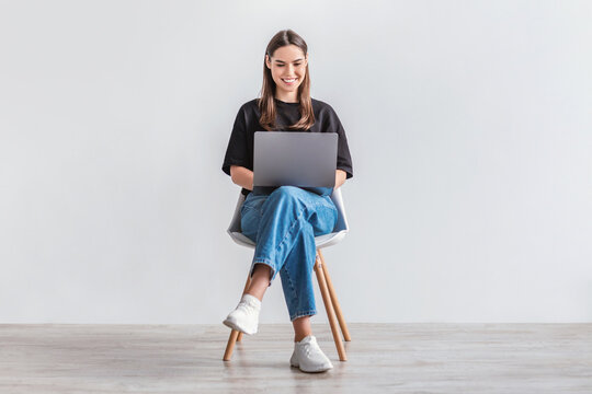 Happy young woman working online, sitting on chair and using laptop against white studio wall, full length