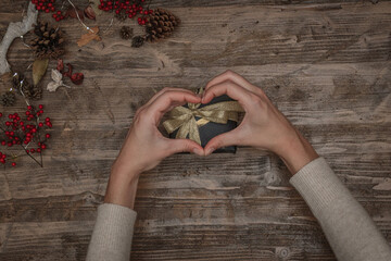 Female hands making a heart shape above a christmas gift on a wooden table with pine cones, berries and led lights