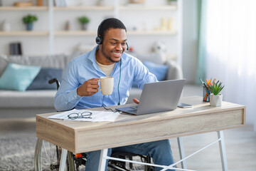 Remote job for disabled people. Black guy in wheelchair wearing headset, using laptop, drinking...