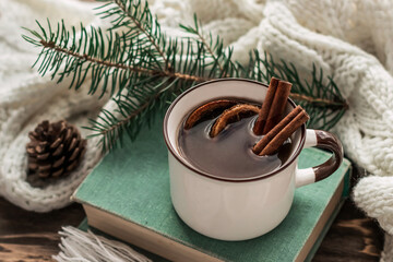 Mug of warming drink with cinnamon sticks on a book on a background of fir branches and cones. Knitted white scarf envelops a mug of hot mulled wine with spices on a wooden background. Cozy winter