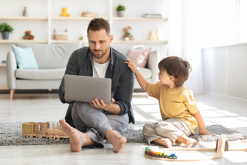 Young concentrated man working on laptop at home, sad little boy touching his shoulder, calling...