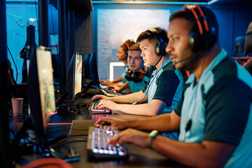 Group of multi-ethnic cybersport players in headsets sitting in front of computers and playing video games online in computer club