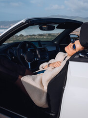 Outdoor lifestyle fashion portrait of stunning girl travelling behind the wheel cabriolet. Stopping and having rest at the ocean. Wearing stylish fur coat, boots, sunglass. Travel by car. Amazing view