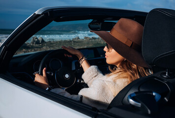 Outdoors lifestyle fashion portrait of stunning young woman driving cabriolet. Driving towards the ocean. Girl travelling behind the wheel. Wearing stylish sheepskin coat, sunglasses, hat. Close up