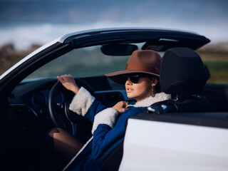 Outdoors lifestyle fashion portrait of stunning young woman driving cabriolet. Girl travelling behind the wheel. Wearing stylish jeans coat, hat, sunglasses. Woman driving. Sensual mood. Road trip