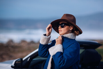 Outdoors lifestyle fashion portrait of stunning young woman travelling behind the wheel convertible. Looking at the ocean, holding hat.  Wearing stylish jeans coat, hat, sunglasses, hat. Travel by car