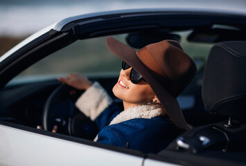 Fototapeta na wymiar Outdoors lifestyle fashion portrait of pretty young woman driving cabriolet. Happy girl smiling behind the wheel. Wearing stylish jeans coat, hat, sunglass. Woman driving. Travel by car. Close up