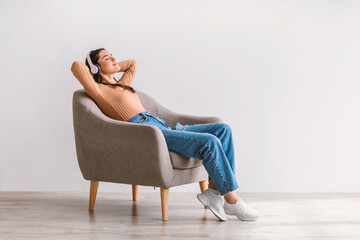 Full length of relaxed young woman listening to music in wireless headphones, resting in armchair...