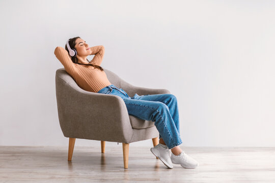 Full length of relaxed young woman listening to music in wireless headphones, resting in armchair against white wall