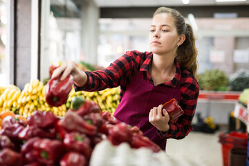 Young positive woman seller lay out ripe pepper on the counter in supermarket