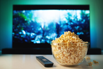 A glass bowl of popcorn and remote control in the background the TV works. Evening cozy watching a movie or TV series at home