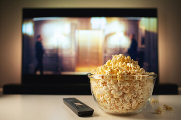 A glass bowl of popcorn and remote control in the background the TV works. Evening cozy watching a...