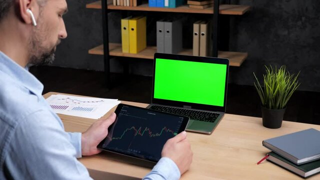 Man stock trader broker looks at tablet display with stock exchange chart analyzes cryptocurrency quotes, laptop with green screen. Businessman freelancer sitting on chair at desk work in office