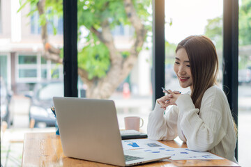 Obraz premium Asian businesswoman working with marketing graphs and laptop in online working concept with financial documents and analytical calculator while sitting in the office.