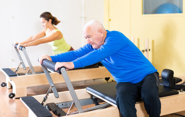 Portrait of elderly man performing set of pilates exercises on reformer during group workout. Active lifestyle and wellness concept of seniors