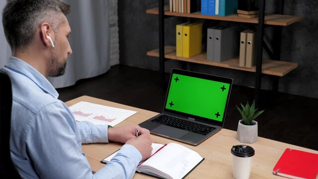 Man professional stock trader broker makes notes in notebook uses laptop with green screen. Businessman writes in notebook looks mock up chroma key computer display sitting on chair at desk in office