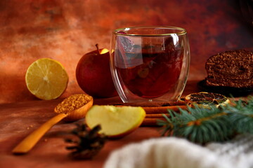 Winter warm drink with cinnamon, star anise and lemon with chocolate roll and spoon of brown sugar. View on table decorated with spices and herbs, apple, fir cones and branch on scarf. Selective focus