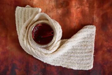 winter warming drink in a glass cup wrapped in a white scarf with a stick of cinnamon, star anise and a dehydrated slice of lemon on brown background. Top view