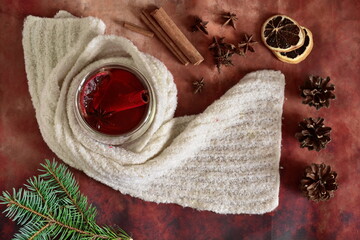 winter warming drink with cinnamon, star anise and lemon in glass cup wrapped in white scarf. Top view on table decorated with spices and herbs, dehydrated slice of lemon, fir cones and branch