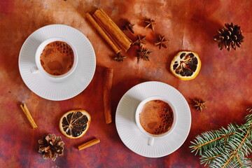 Winter warming drink hot liquid cocoa chocolate with foam. Top view on brown table decorated with spices and herbs, cinnamon sticks and star anise, dehydrated slice of lemon, fir cones and branch
