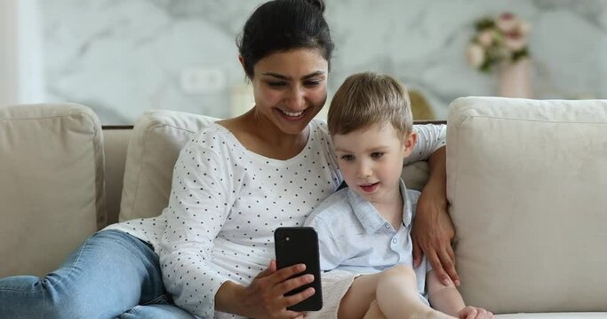 Happy young mom sit on sofa hug cute little boy hold phone watch funny cartoon online on gadget screen. Smiling family of Indian stepmother European son use modern cell talk by video call