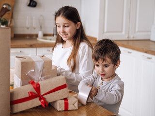 Cute toddler caucasian boy in pajama (in focus)sitting in bright kitchen and packing Christmas boxes with sister