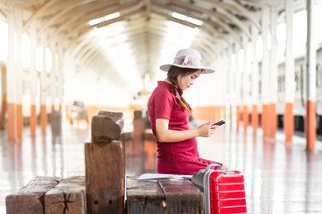 Asian woman pregnant in red dress carrying red luggage and look at the Smartphone with a red suitcase at railway station travel,traveler with backpack in summer Holiday concept Thailand