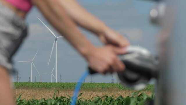 Windmills rotate in corn field under cloudy sky. Young woman in shorts connects plug to white electric car at charging station closeup