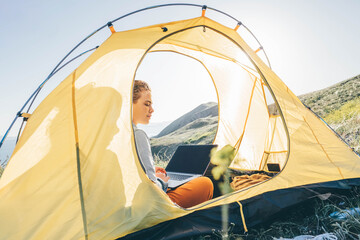 Young woman in yellow tent and types on grey laptop on sunny day.