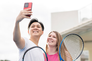 Hombre asiático y una mujer caucásica tomando un selfie después de jugar al tenis con raquetas...